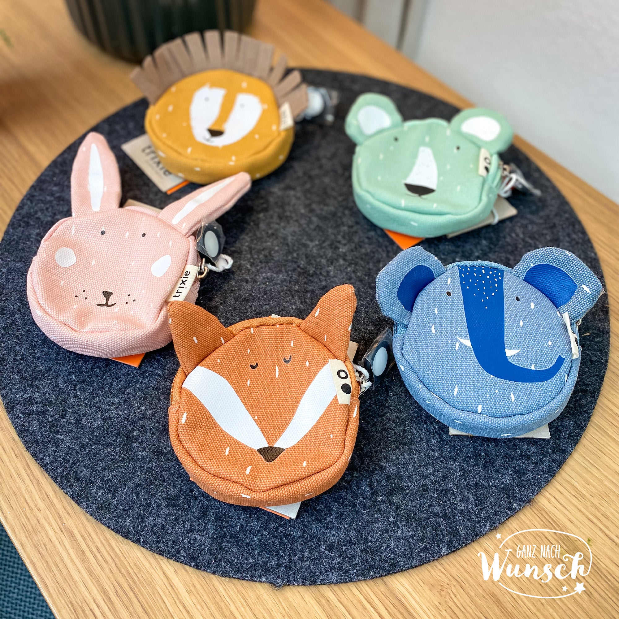 Fünf farbenfrohe Kindergeldbörsen in Tierdesigns: rosa Hase, gelber Löwe, grüner Bär, orange Fuchs, blauer Elefant. Runde Stoffbörsen mit niedlichen Gesichtern auf grauem Filzuntersetzer. Perfekt für Kinder, praktisch und spielerisch zugleich.