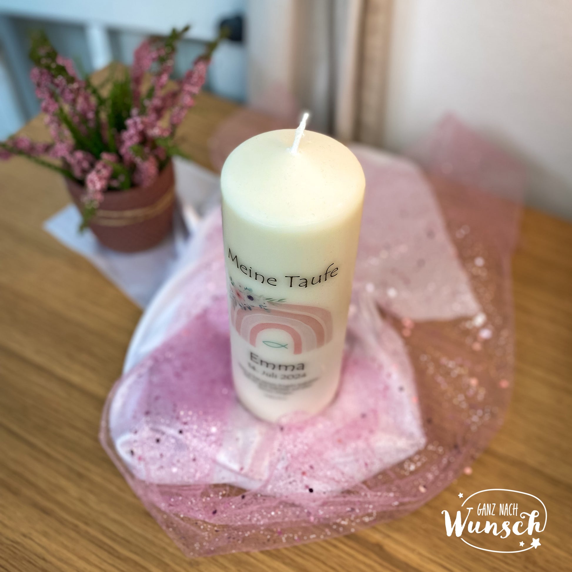 Eine personalisierte Taufkerze mit einem pastellfarbenen Regenbogenmotiv, dem Namen, dem Taufdatum und einem christlichen Fischsymbol. Umgeben von rosa Tüll, festliche und friedliche Atmosphäre, mit violetter Topfpflanze im Hintergrund.