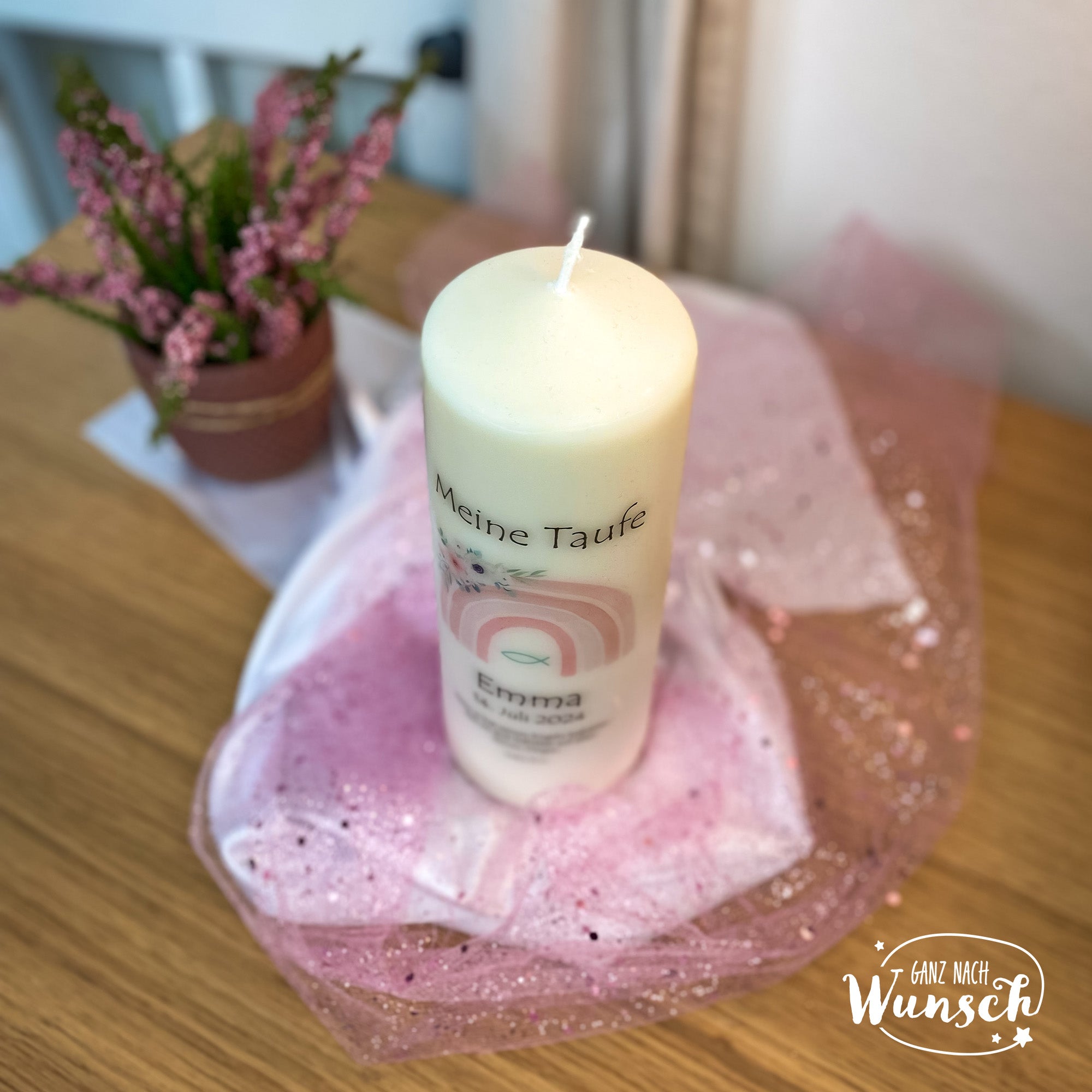 Eine personalisierte Taufkerze mit einem pastellfarbenen Regenbogenmotiv, dem Namen, dem Taufdatum und einem christlichen Fischsymbol. Umgeben von rosa Tüll, festliche und friedliche Atmosphäre, mit violetter Topfpflanze im Hintergrund.