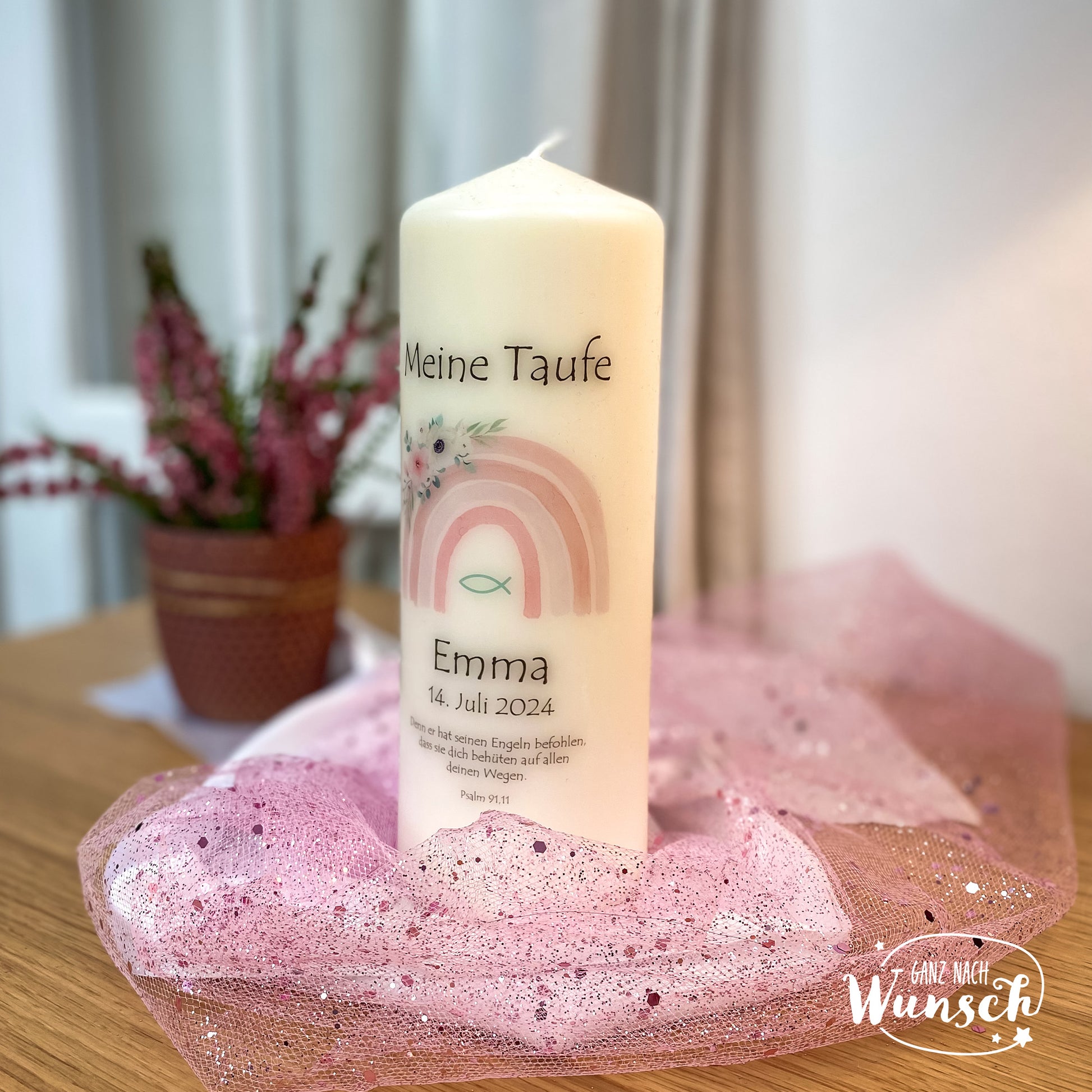 Eine personalisierte Taufkerze mit einem pastellfarbenen Regenbogenmotiv, dem Namen, dem Taufdatum und einem christlichen Fischsymbol. Umgeben von rosa Tüll, festliche und friedliche Atmosphäre, mit violetter Topfpflanze im Hintergrund.